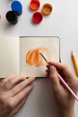 An artist begins painting a pumpkin sketch in a notebook using bright orange gouache. Open jars of paint are nearby, illustrating the process of creating art with vibrant colors. clipart