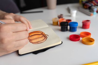 Sanatçı balkabağı resimlerine son rötuşlarını dikkatlice gouache boyaları kullanarak uygular. Canlı renklerin olduğu kavanozlar masanın üzerine yayılıyor ve yaratıcı süreci gösteriyor..