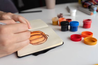 Sanatçı balkabağı resimlerine son rötuşlarını dikkatlice gouache boyaları kullanarak uygular. Canlı renklerin olduğu kavanozlar masanın üzerine yayılıyor ve yaratıcı süreci gösteriyor..