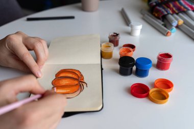 Sanatçı balkabağı resimlerine son rötuşlarını dikkatlice gouache boyaları kullanarak uygular. Canlı renklerin olduğu kavanozlar masanın üzerine yayılıyor ve yaratıcı süreci gösteriyor..