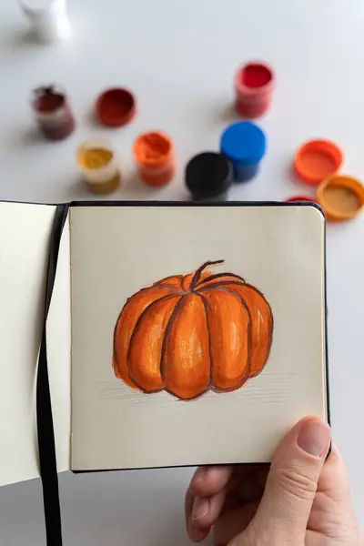 stock image Close-up of an artist mixing orange gouache paint to add details to a pumpkin illustration. Various paint jars are spread on the table, showing the creative process in action.