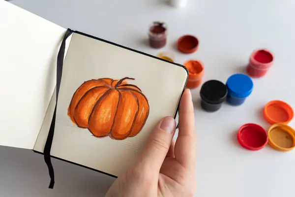 stock image Close-up of an artist mixing orange gouache paint to add details to a pumpkin illustration. Various paint jars are spread on the table, showing the creative process in action.