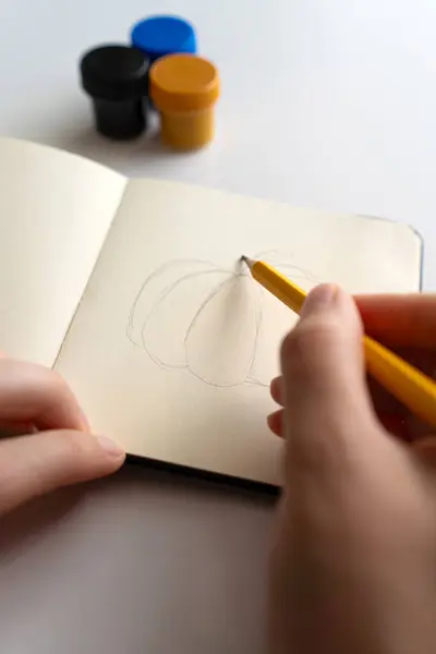 stock image An artist begins sketching a pumpkin with a pencil in a notebook. Three paint jars are placed nearby, illustrating the creative process from the initial sketch to the finished gouache painting.