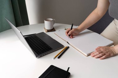 Bir dizüstü bilgisayar, kahve fincanı, defter, kalem ve kalemle beyaz bir masanın üzerine kurulmuş temiz ve minimalist bir ofis. Üretkenlik, uzak çalışma ve yaratıcı planlama için mükemmel..