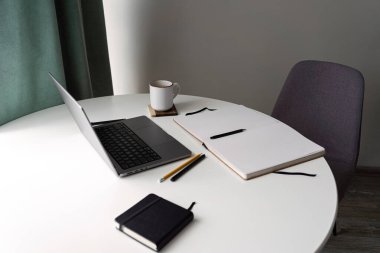 Bir dizüstü bilgisayar, kahve fincanı, defter, kalem ve kalemle beyaz bir masanın üzerine kurulmuş temiz ve minimalist bir ofis. Üretkenlik, uzak çalışma ve yaratıcı planlama için mükemmel..