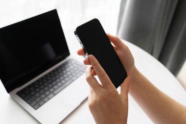 Arka planda dizüstü bilgisayarı olan bir akıllı telefonu tutan ellerin yakın çekimi. Ev ofis ortamında teknoloji, iletişim, uzak çalışma ve dijital yaşam tarzı kavramları için idealdir..