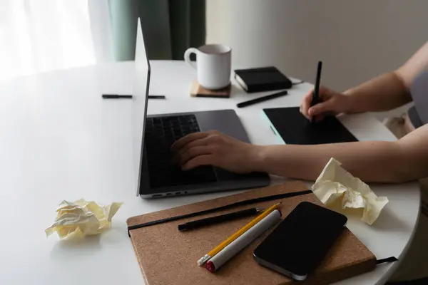 stock image A side view of a creative workspace featuring a laptop, notebook, drawing tablet, and coffee. Crumpled paper symbolizes brainstorming and unsuccessful ideas during the creative process.