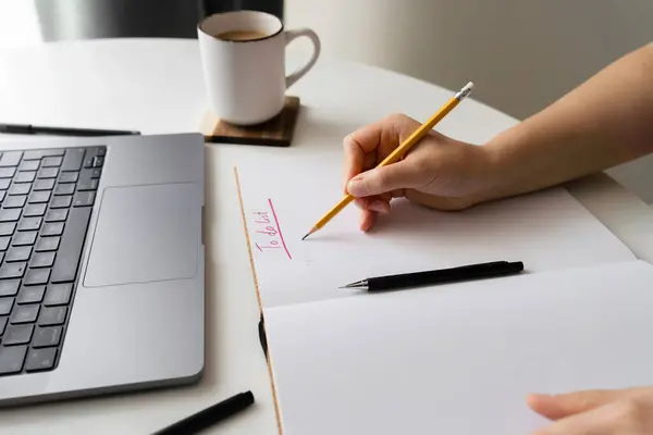 stock image Side view. A person writing a to-do list in a notebook with a laptop and stationery on a desk. Ideal for concepts of productivity, planning, organization, and remote work setup.