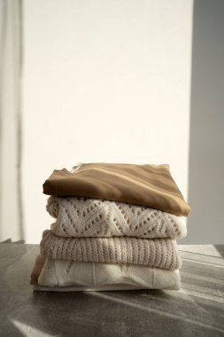 Yumuşak bej tonlarda düzgünce katlanmış, örülmüş bir kazak yığını bir yatağın üzerine konur. Güneş ışığı yünün dokusunu güçlendirir ve sıcak ve davetkar bir atmosfer yaratır. Terfi için mükemmel.