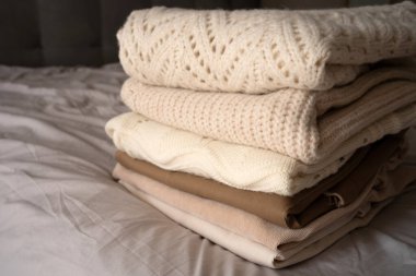 Yumuşak, nötr tonlarda düzgün örülü kazaklar bir yatakta oturur. Süveter dokuları ve desenleri sıcaklığı ve konforu iletir. Sürdürülebilir moda ve ikinci el kıyafetleri desteklemek için mükemmeldir.