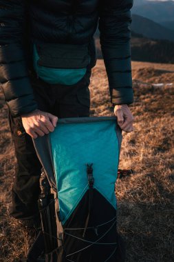 Altın saat boyunca otlak bir dağ sırtında yürüyüş malzemelerini büyük bir sırt çantasına koyan bir yürüyüşçünün yakın çekimi. Batan güneş sıcacık bir ışık saçıyor, açık hava macera takımlarını vurguluyor..