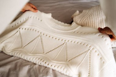 Bir kadın, beyaz örgü bir kazağı yanında sıcak, nötr renkli giysiler yığını olan bir yatakta düzgünce katlıyor. Sahne, minimalist modayı, rahat titreşimleri ve sürdürülebilir yaşamı yansıtıyor. Ev için mükemmel.
