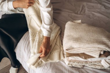 Bir kadın, beyaz örgü bir kazağı yanında sıcak, nötr renkli giysiler yığını olan bir yatakta düzgünce katlıyor. Sahne, minimalist modayı, rahat titreşimleri ve sürdürülebilir yaşamı yansıtıyor. Ev için mükemmel.