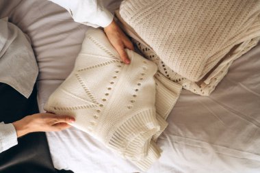 Bir kadın, beyaz örgü bir kazağı yanında sıcak, nötr renkli giysiler yığını olan bir yatakta düzgünce katlıyor. Sahne, minimalist modayı, rahat titreşimleri ve sürdürülebilir yaşamı yansıtıyor. Ev için mükemmel.