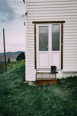 Basamakta bir çift yıpranmış botun olduğu basit bir köy evi girişi. Sahne, arka planda dalgalı tepeler ve dağlarla yaşayan kırsal kesimin sessiz basitliğini yakalıyor.