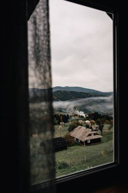 Uzak evler, sisli dağlar ve huzurlu bir kırsal alan içeren sakin bir köy manzarası bir pencereden izleniyor. Yumuşak sis ve kırsal cazibe huzuru ve sadeliği çağrıştırıyor.