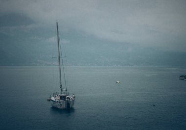 Yalnız bir yelkenli gemi bulutlu bir gökyüzünün altındaki yüksek dağlarla çerçevelenmiş sakin sularda yüzer. Dramatik manzara, açık denizde barışçıl ama maceralı yolculuğu vurgular..