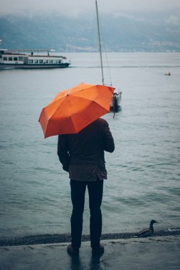 Canlı turuncu bir şemsiyenin altında, gölün sakin sularında bir insan dururken, botlar ve bir ördek, yağmurlu ve dingin bir ortama renk katıyor. Parlak şemsiye, sisli havada figürü öne çıkarır.