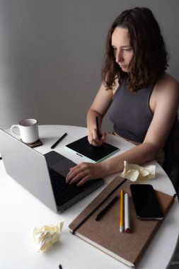 Bir kadın hem bir dizüstü bilgisayar hem de dijital tablet kullanarak ev ofisindeki işine konsantre olur. Masadaki çeşitli maddeler, bir defter ve buruşuk kağıt da dahil, yoğun bir çalışma oturumu öneriyor.