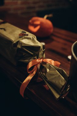 A wrapped gift with a shiny orange ribbon sits on a wooden table, capturing the cozy, festive atmosphere of Halloween decor with soft lighting and a blurred pumpkin in the background. clipart