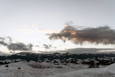 Dramatik bulutlarla dolu uçsuz bucaksız gökyüzünün altındaki karla kaplı dağların resimli görüntüsü. Akşam ışığı manzaraya yumuşak bir parıltı yayar, huzurlu ve huzurlu bir atmosfer yaratır..