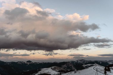 Dramatik bulutlarla dolu uçsuz bucaksız gökyüzünün altındaki karla kaplı dağların resimli görüntüsü. Akşam ışığı manzaraya yumuşak bir parıltı yayar, huzurlu ve huzurlu bir atmosfer yaratır..