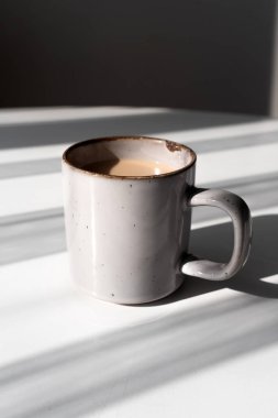 Kahve ya da çayla dolu seramik bir kupa beyaz bir yüzeye dramatik bir gölge düşürüyor. Sabah rutinleri, minimalizm, rahatlama ve sakin anlar için mükemmel kavramlardır..