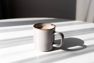 Kahve ya da çayla dolu seramik bir kupa beyaz bir yüzeye dramatik bir gölge düşürüyor. Sabah rutinleri, minimalizm, rahatlama ve sakin anlar için mükemmel kavramlardır..