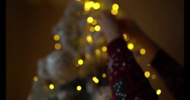 Bayram süveteri giyen kadın ellerinin yakın çekimi, rahat bir tatil ortamında parıldayan sarı ışıklarla bir Noel ağacına bir süs asmak. Noel, kış, tatil temaları için mükemmel.