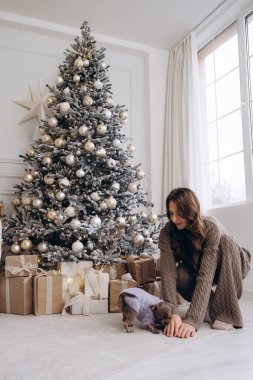 Güzel dekore edilmiş, altın ve beyaz süslemeli ve güzelce paketlenmiş bir Noel ağacının olduğu samimi bir Noel sahnesi. Örgü kıyafetli bir kadın ağacın yanında diz çöküp onunla oynuyor.