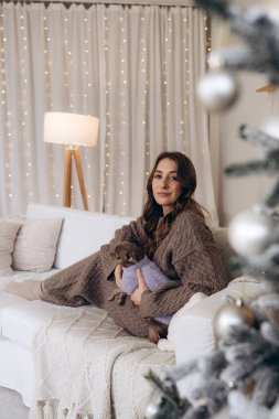 Sıcak bir kanepede oturan, kalın örgü bir elbise giyen bir kadın, çuvava köpeğini mor bir kazak içinde tutuyor. Sahne güzel bir şekilde süslenmiş Noel süsleri, parlayan peri ışıkları, ve