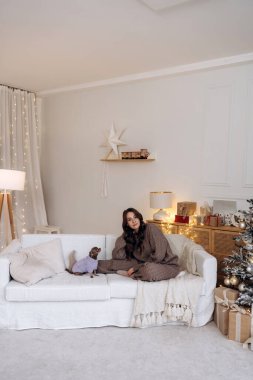 Köpeğiyle beyaz bir kanepede oturan rahat bir kadın sahnesi. Etrafı zarif Noel süsleri ve şenlikli bir iç mekanda parlayan ışıklarla çevrili..