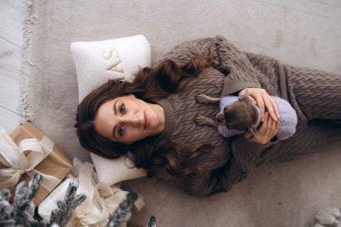 A young woman holding her dog while lying on a Christmas-themed carpet surrounded by gifts and decorations, enjoying a cozy festive morning. clipart