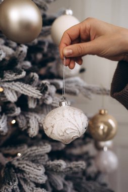 A close-up of a hand in a cozy knitted sweater placing a white ornament on a frosted Christmas tree with golden decorations, creating a festive and elegant atmosphere. clipart