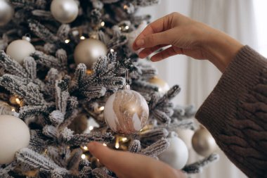 Sıcak örülmüş bir süveterin içinde, altın süslemeli beyaz bir Noel ağacına beyaz bir süs takan bir elin yakın çekimi, şenlikli ve zarif bir atmosfer yaratıyor..