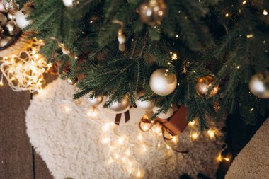 Altın ve bronz Noel süsleriyle dolu bir hasır sepetin yakın çekimi, şenlikli bir ağacın yanında ve parlayan peri ışıklarının yanında. Mevsimlik dekorasyon için sıcak bir tatil ortamı.