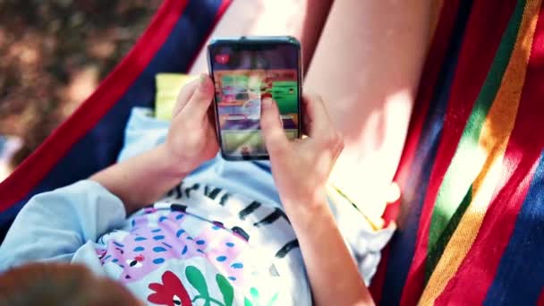 好奇的小女孩躺在自然的吊床上玩游戏 一边用手机 一个小孩手里拿着一部手机 在大自然中玩游戏 儿童的技术成瘾概念 儿童游戏成瘾 — 图库视频影像
