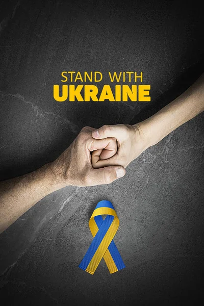 stock image hand holding tight hand of child with yellow and blue ribbon on marble, with words stand with Ukraine.