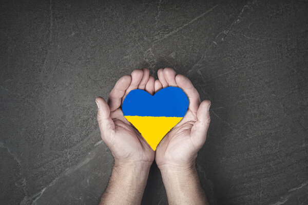 male hands hold painted yellow and blue heart on grey marble background. concept needs help and support, truth will win