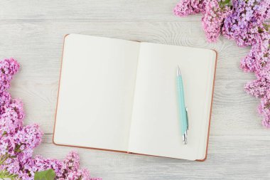 Open diary with a pen and lilac flowers on a white wooden background. Top view.
