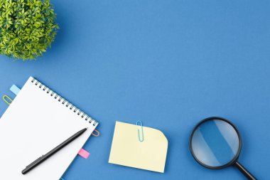 Kağıt ataçlarından kitap ayracı, not kağıtları, kalem, büyüteç ve mavi izole arka planda mühürlü yaprakları olan ev bitkisi içeren sarmal defter. Ofis kavramı. Üst görünüm.