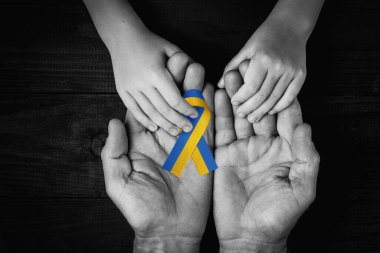 male hands hold childrens on dark wooden background with yellow and blue ribbon. black and white color. concept needs help and support, truth will win