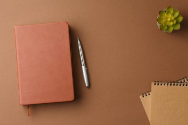 Kalemli el işi kitabı, sarmal defterler, kahverengi arka planda yeşil yapraklı ev bitkisi. Organizatör çalışma masasında. Üst görünüm.