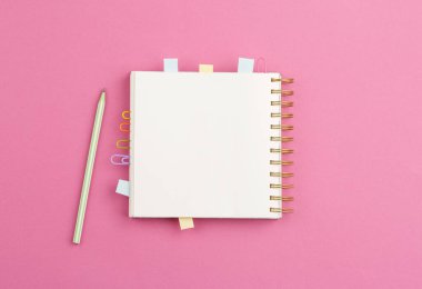 Kağıt ataçlarının yer imlerini içeren spiral bir defter aç ve notlar için yapraklar, pembe arka planda bir kalem. Üst görünüm.