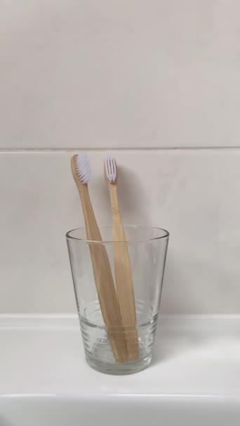 女性の手はガラスカップから竹の歯ブラシを取ります 垂直ビデオ バスルーム内の生分解性衛生製品 — ストック動画