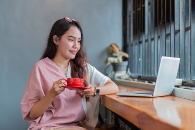 Genç Asyalı kadın elinde bir fincan kahve tutuyor ve kırıyor. Kafedeki güzel Asyalı iş kadını. Laptop kullanıyor, kahve içiyor ve gülümsüyor.