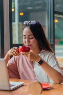 Genç Asyalı kadın elinde bir fincan kahve tutuyor ve kırıyor. Kafedeki güzel Asyalı iş kadını. Laptop kullanıyor, kahve içiyor ve gülümsüyor.