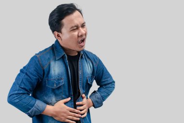 Genç Asyalı bir adamın midesi ağrıyor ya da hazımsızlık çekiyor ve karnını izole edilmiş beyaz arka plana bastırıyor..