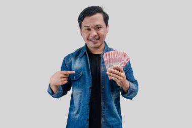 Elinde kağıt para tutarken ve kameraya bakarak parayı işaret ederken mutlu bir yüz ifadesi gösteren genç Asyalı adam.