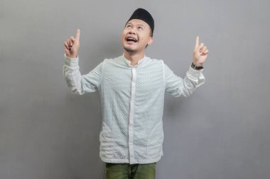 Mutlu Asyalı Müslüman adam tanıtımı fotokopi alanını işaret ederek oruç ayının gölgeleriyle koko gömlek ve göğüs kafesi giyerek gri bir arkaplanda izole edilmiş bir halkayı işaret ederek tavsiye ediyor.